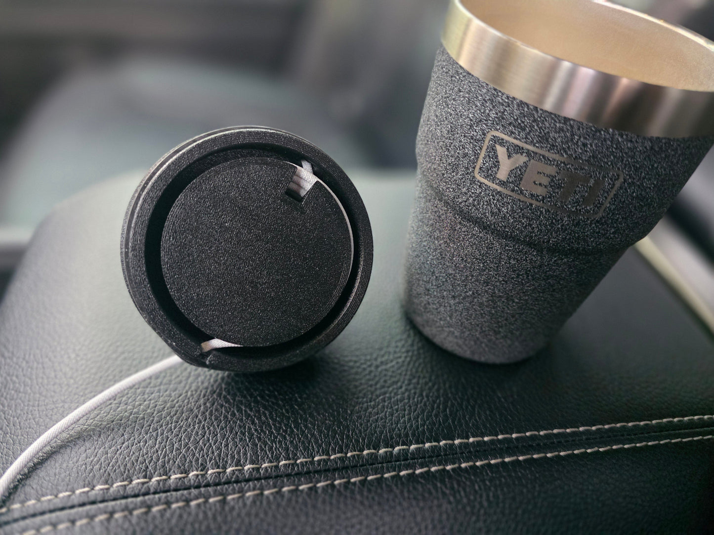 Underside of the black MagSafe stand lid, showing the charging cable coming up through a slot in the lid and coiled around the built-in cable channel, next to an open YETI cup