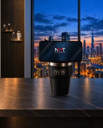 Phone docked on an NMT replacement lid fitted to a black YETI cup on a desk overlooking a city at dusk, with YETI and Stanley cups on wall ledges behind.