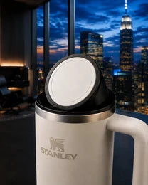 Black NMT replacement lid and charging puck on a white Stanley Quencher, beside a city window.